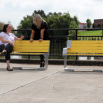 Two women sit and talk on a yellow bench near a pond, with another empty bench beside them and a college building visible in the background.