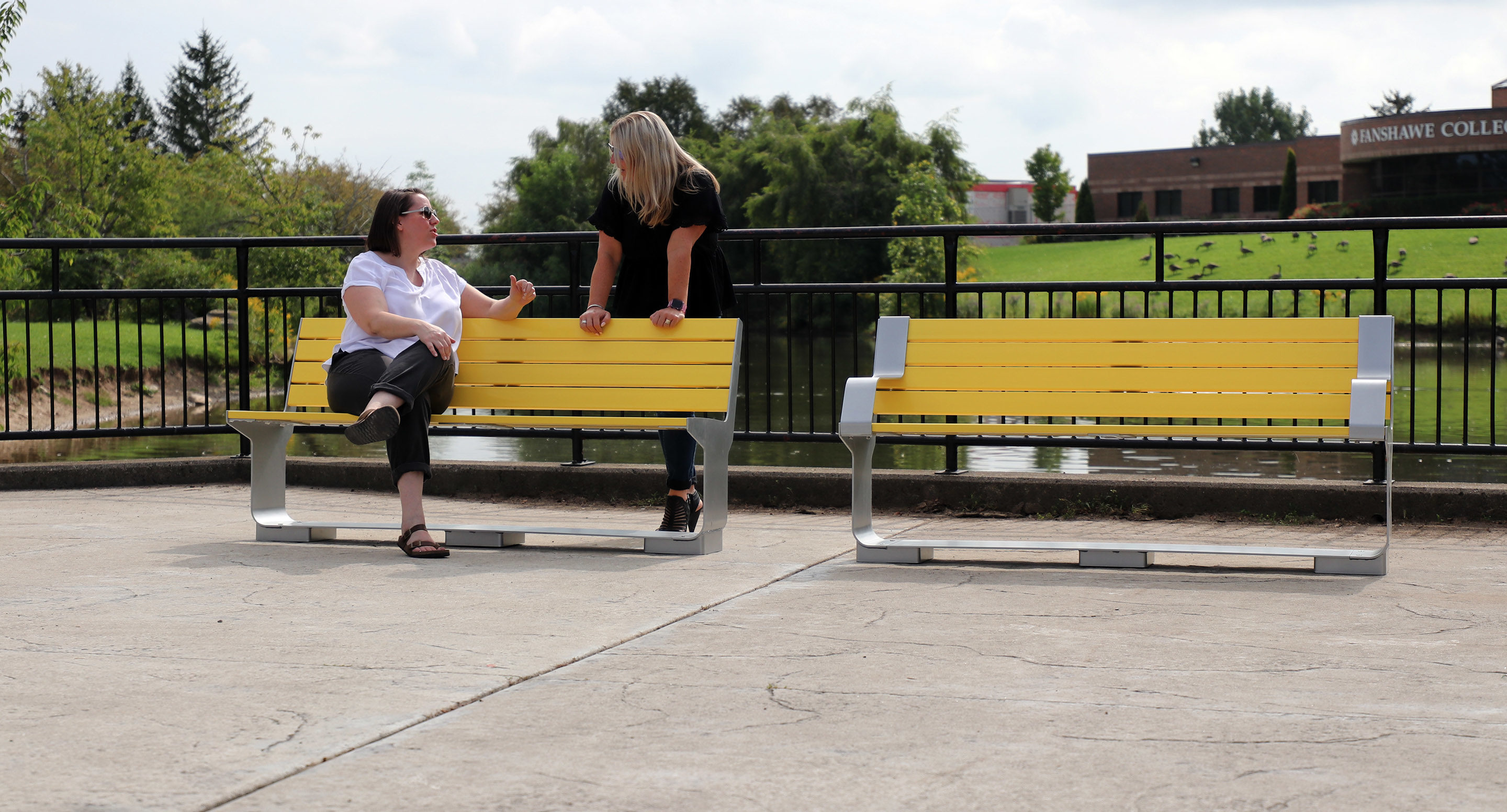 maglin_mbe-3000-backed-benches-2 Two women sit and talk on a yellow bench near a pond, with another empty bench beside them and a college building visible in the background.