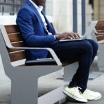 A person in a blue blazer and white sneakers is sitting on a bench outdoors, using a laptop on their lap. Another bench and a smartphone are visible nearby.