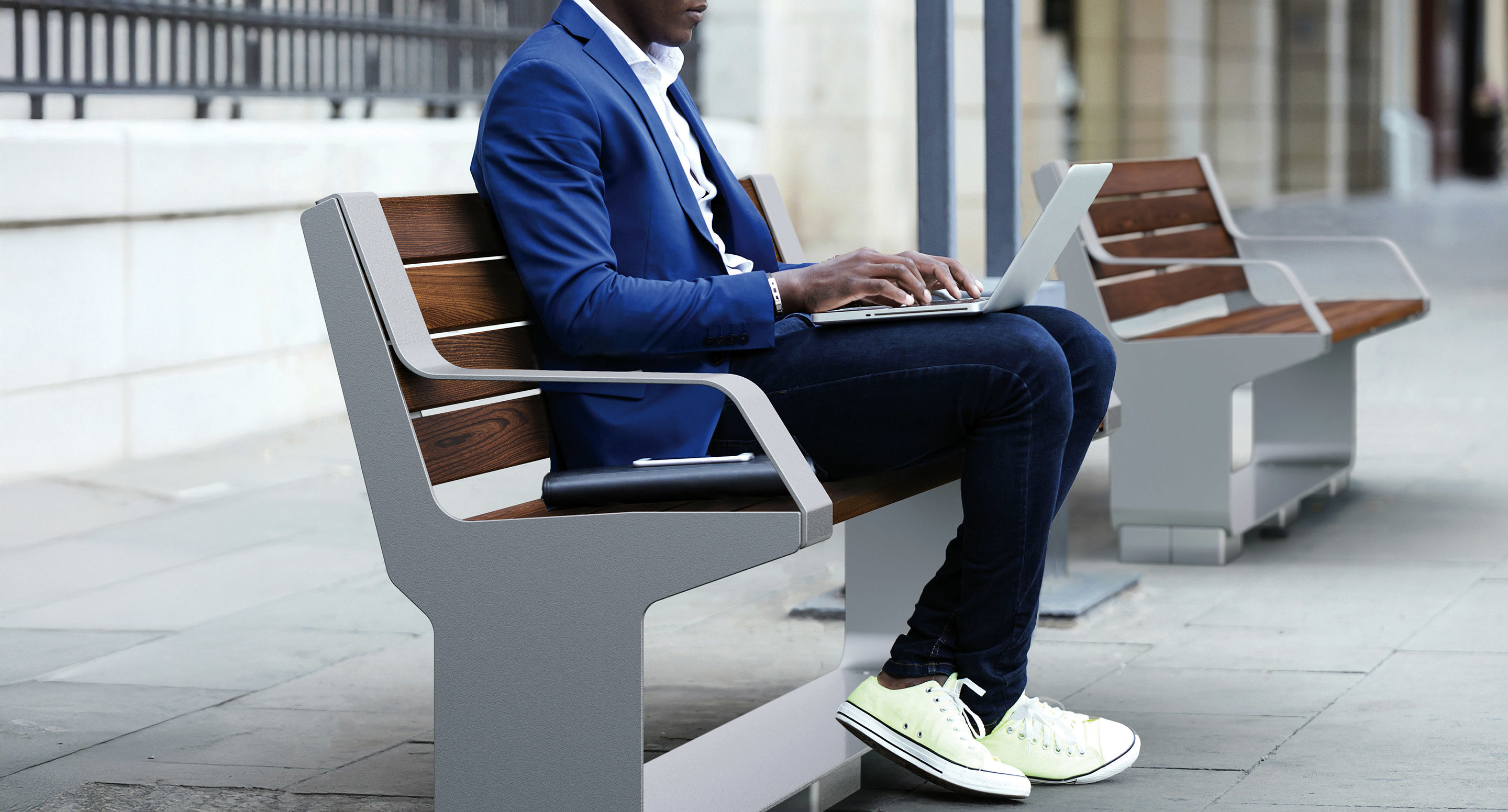 maglin_mbe-3000-backed-benches-3 A person in a blue blazer and white sneakers is sitting on a bench outdoors, using a laptop on their lap. Another bench and a smartphone are visible nearby.