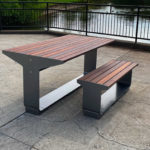 A modern outdoor picnic table with an attached bench, made of metal and wood, is placed on a paved surface near a pond with a black metal railing in the background.
