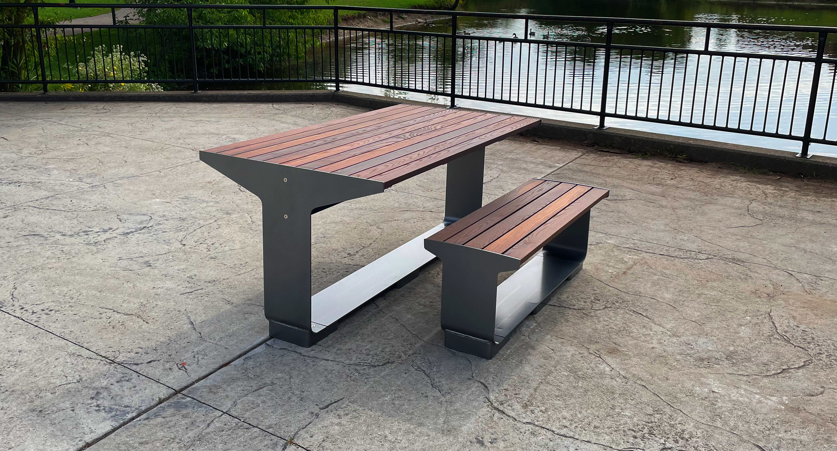 maglin_mbe-3000-backless-benches-1 A modern outdoor picnic table with an attached bench, made of metal and wood, is placed on a paved surface near a pond with a black metal railing in the background.