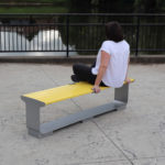 A person sits on a yellow bench facing a pond, with their back to the camera, near a metal railing on a paved surface.