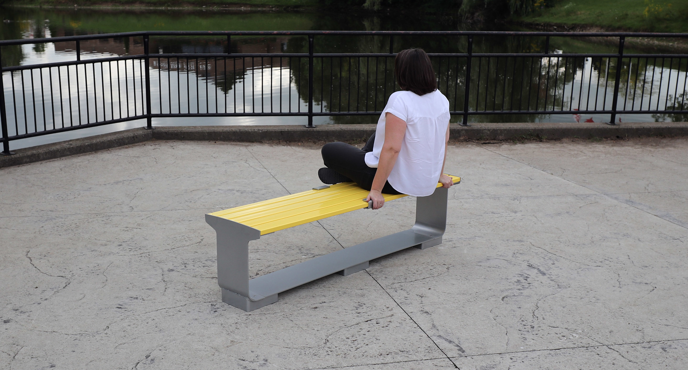 maglin_mbe-3000-backless-benches-2 A person sits on a yellow bench facing a pond, with their back to the camera, near a metal railing on a paved surface.