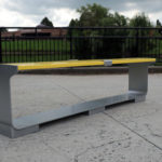 A modern bench with a yellow top and gray metal sides is placed on a paved outdoor area near a black metal railing, with trees and a building in the background.