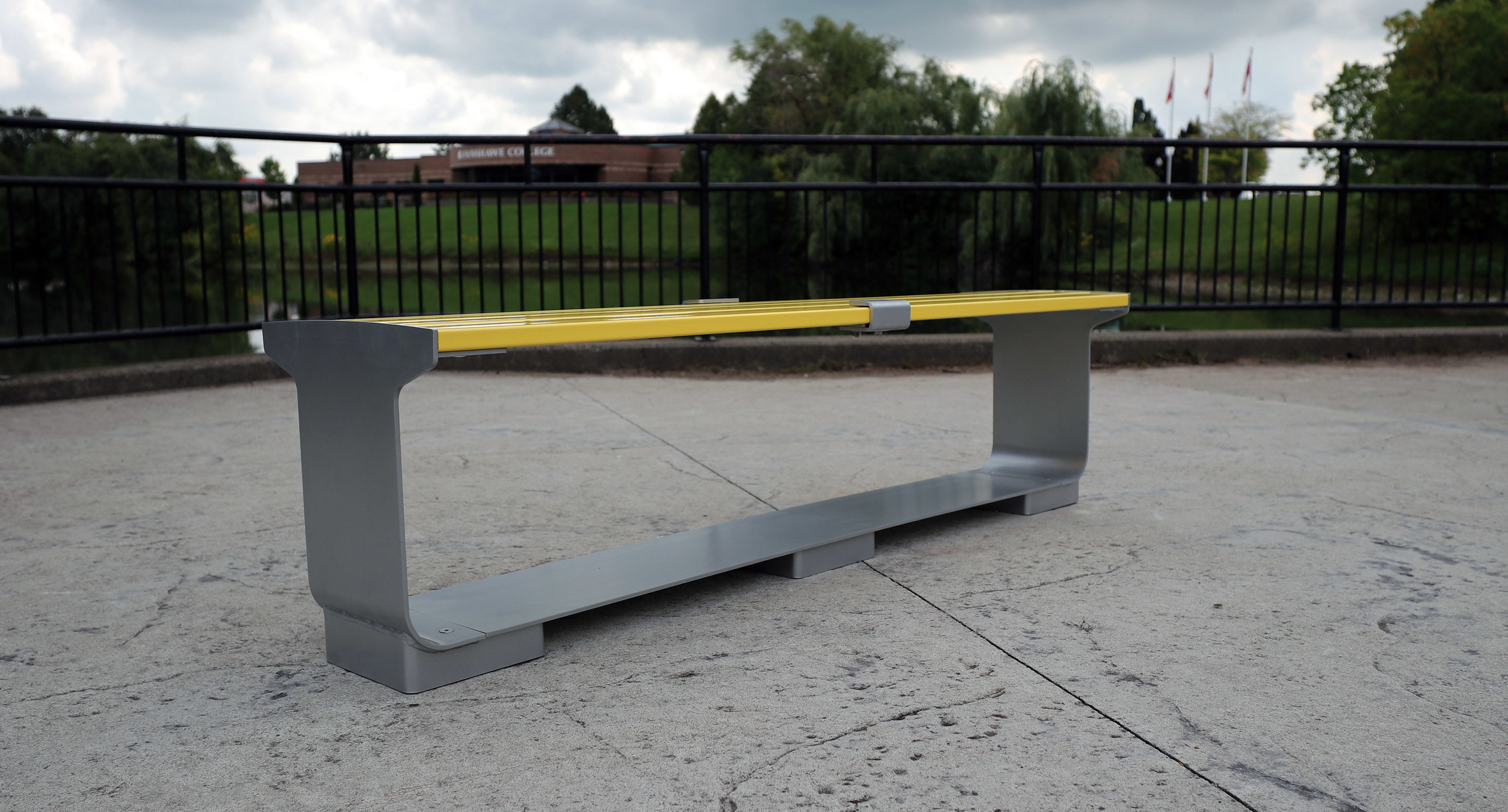 maglin_mbe-3000-backless-benches-3 A modern bench with a yellow top and gray metal sides is placed on a paved outdoor area near a black metal railing, with trees and a building in the background.