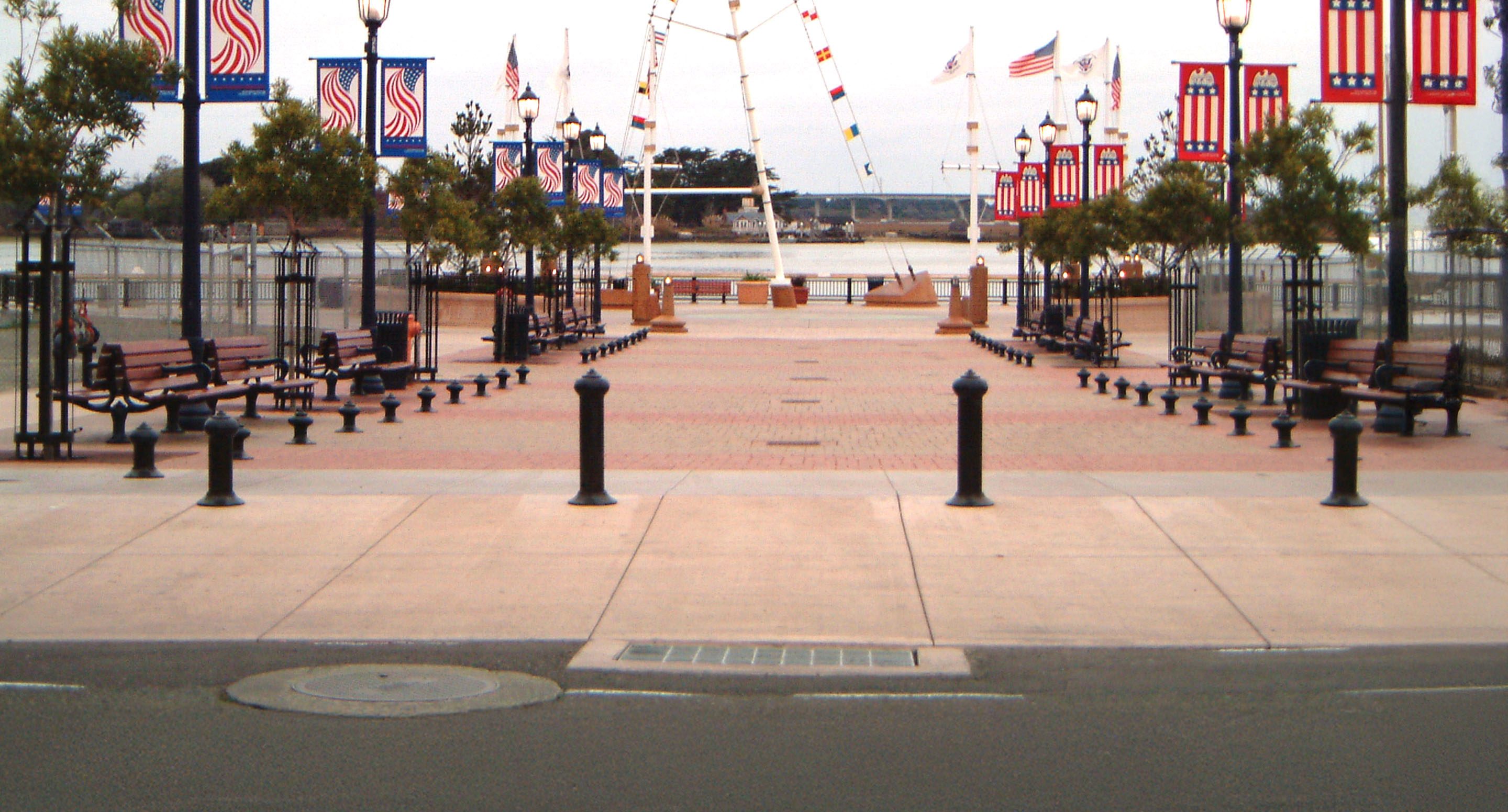 maglin_mbo-0100-bollards-1 Une allée pavée bordée de bancs, d'arbres, de lampadaires et de bannières patriotiques mène à un plan d'eau où flottent plusieurs drapeaux américains.