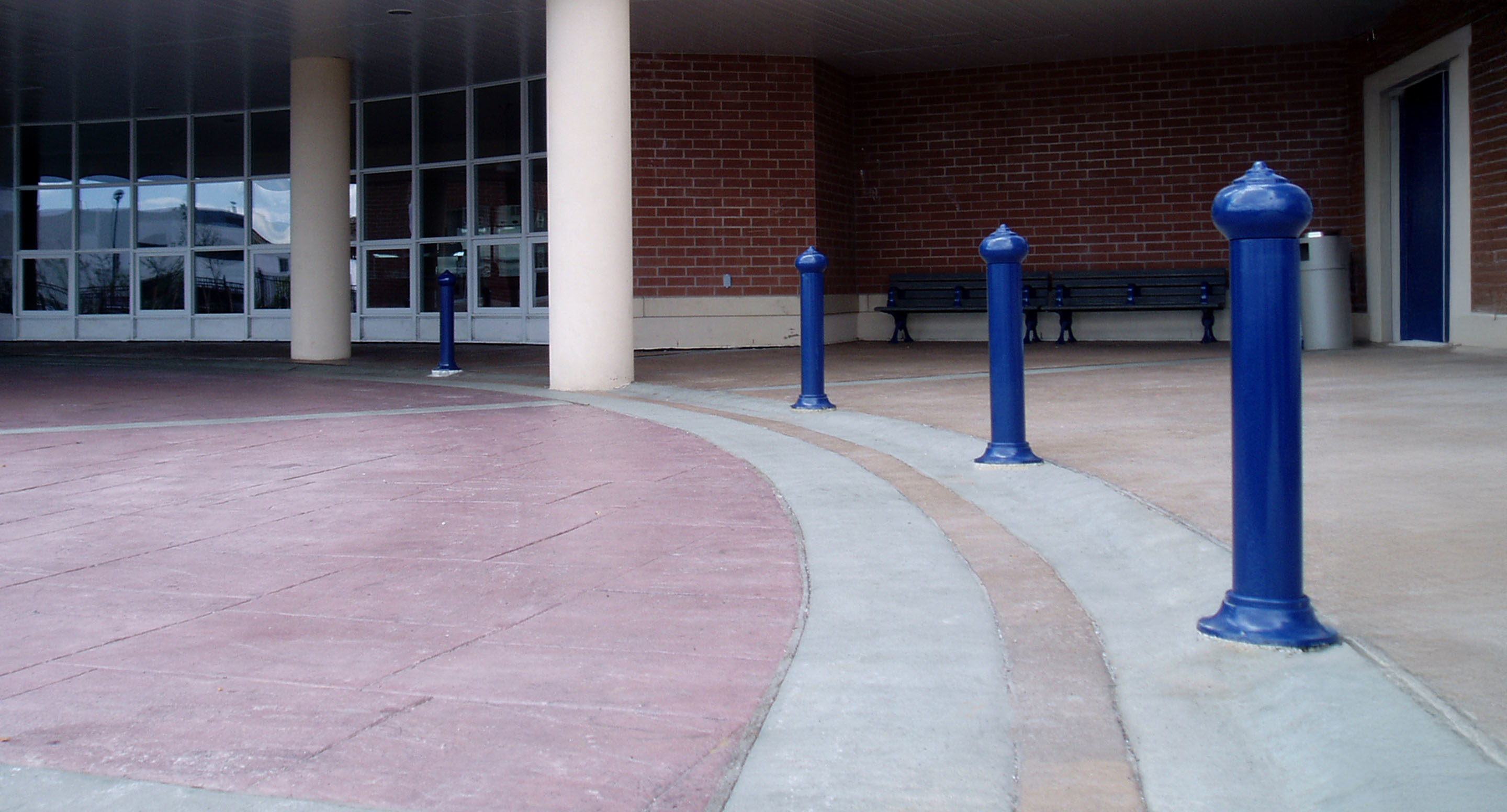 maglin_mbo-0100-bollards-2 Des bornes bleues bordent une allée courbe devant l'entrée d'un bâtiment avec des murs en briques, de grandes fenêtres et des bancs à l'arrière-plan.