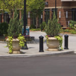 Une personne est assise sur un banc dans la cour d'un centre commercial avec, en arrière-plan, de grandes jardinières, des bornes, des arbres et des devantures de magasins.