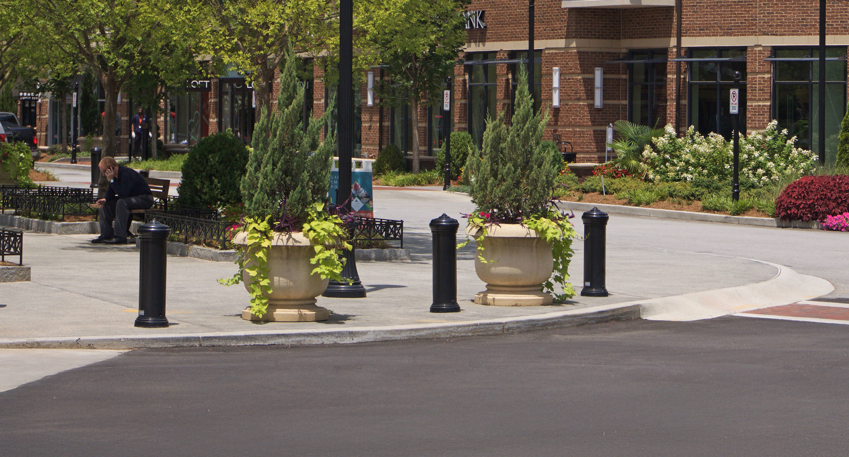 maglin_mbo-0200-bollards-2 Une personne est assise sur un banc dans la cour d'un centre commercial avec, en arrière-plan, de grandes jardinières, des bornes, des arbres et des devantures de magasins.