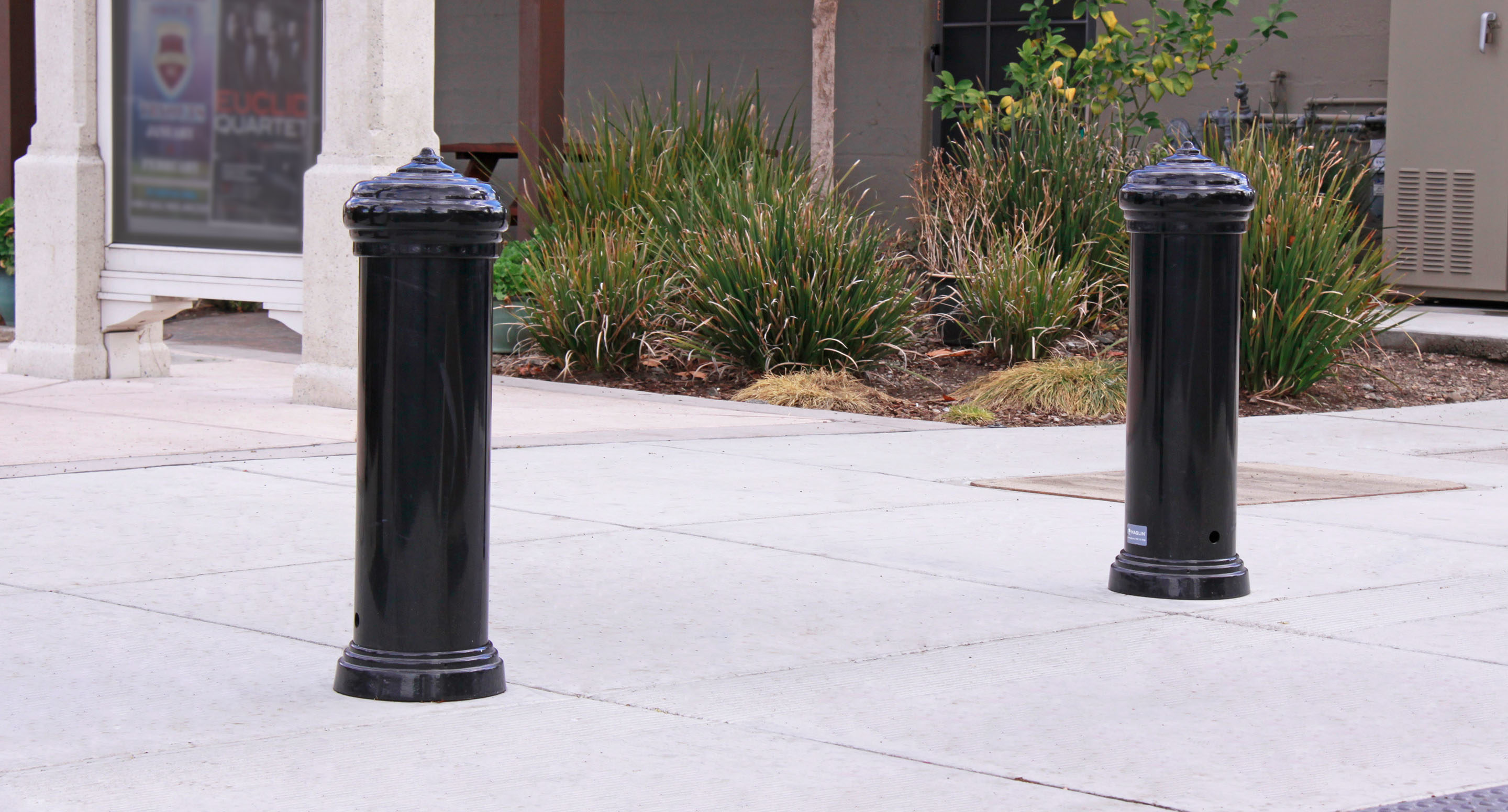 maglin_mbo-0200-bollards-3 Deux bornes métalliques noires sont installées sur un trottoir devant un espace paysager avec des buissons et un mur d'enceinte à l'arrière-plan.