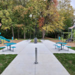 Aire de jeux extérieure avec deux structures de jeux, des balançoires, des tables de pique-nique et une allée en béton, entourée de gazon et d'arbres au feuillage vert et automnal.