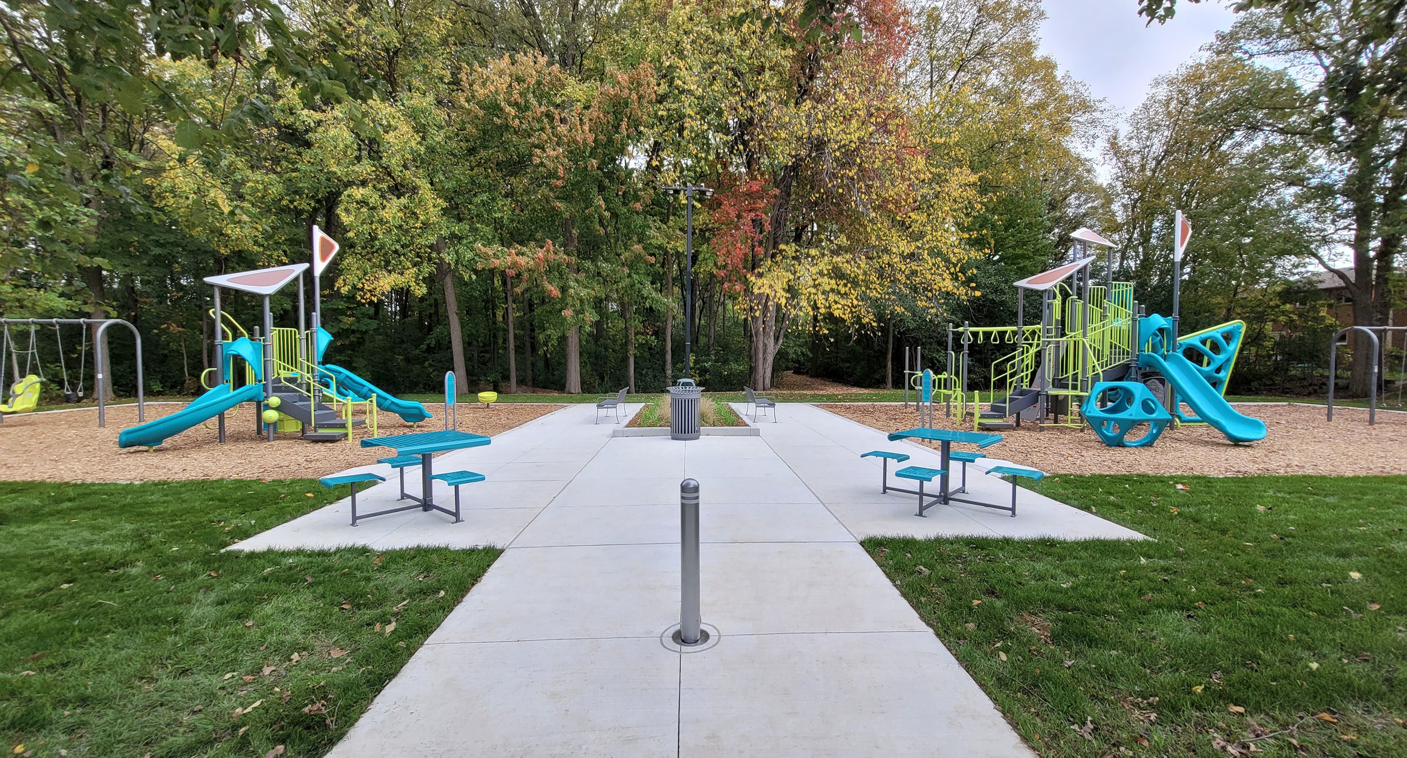 maglin_mbo-0500-bollards-1 Aire de jeux extérieure avec deux structures de jeux, des balançoires, des tables de pique-nique et une allée en béton, entourée de gazon et d'arbres au feuillage vert et automnal.