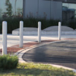 Cinq bornes argentées bordent un trottoir incurvé à côté d'un bâtiment aux grandes baies vitrées, d'un aménagement paysager et d'une route pavée au premier plan.
