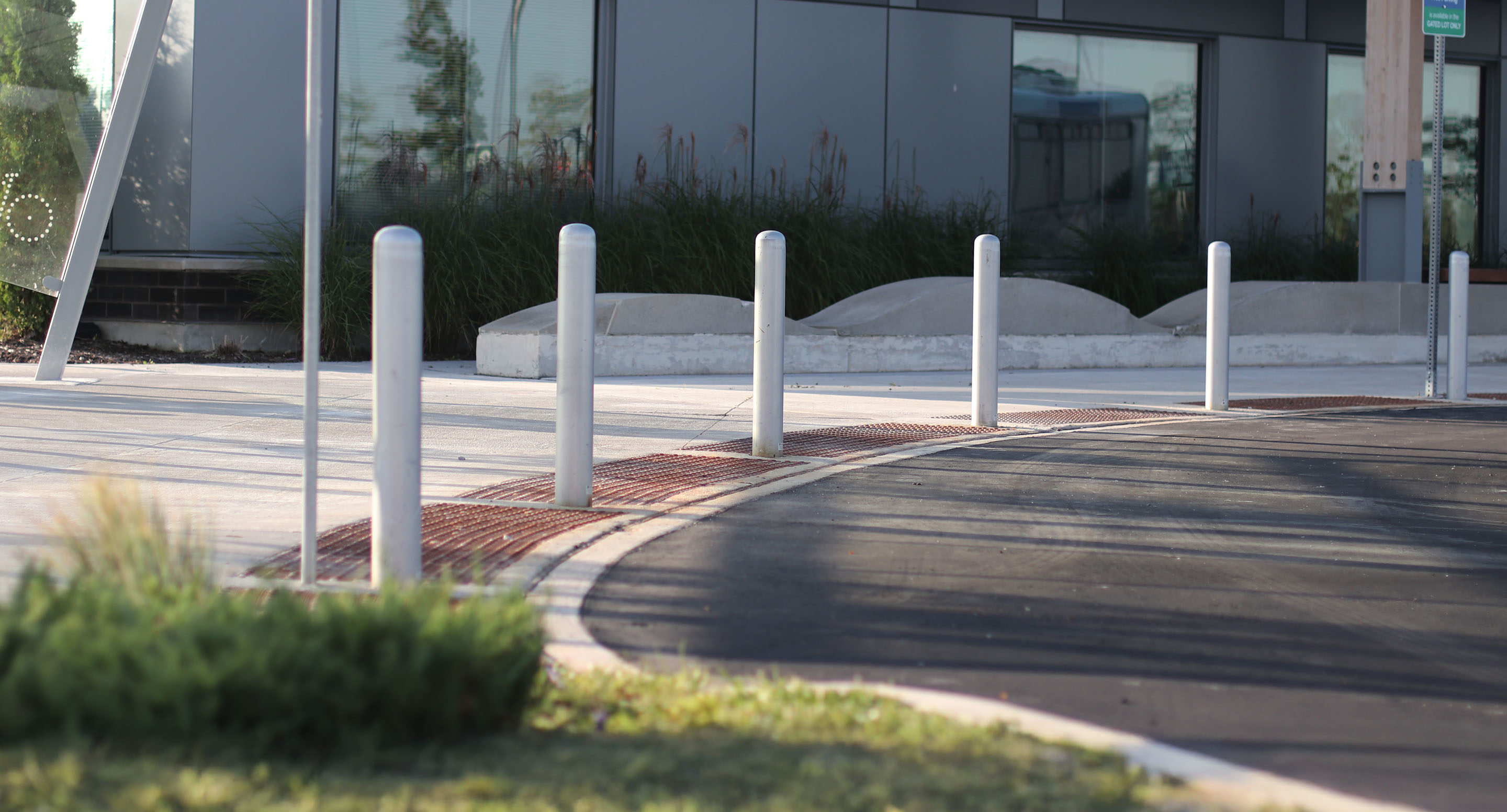 maglin_mbo-0500-bollards-2 Cinq bornes argentées bordent un trottoir incurvé à côté d'un bâtiment aux grandes baies vitrées, d'un aménagement paysager et d'une route pavée au premier plan.
