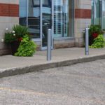 Quatre bornes métalliques et deux grandes jardinières avec des plantes vertes et rouges bordent un trottoir devant une vitrine, à côté d'une voiture garée dans un parking.