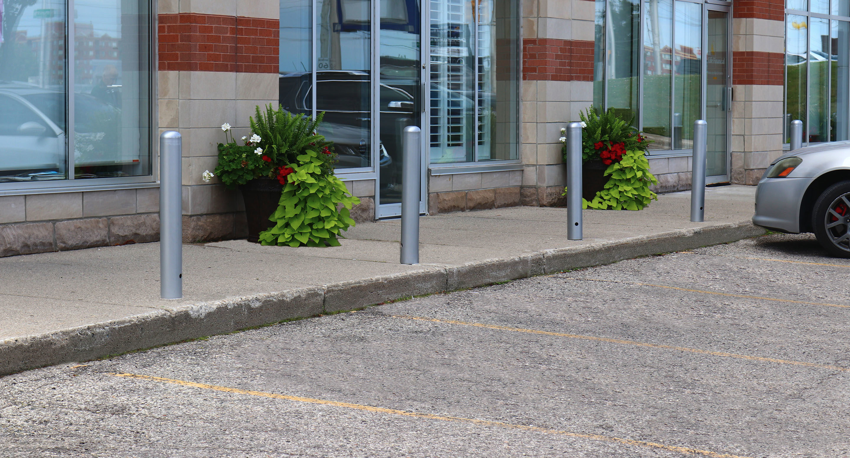 maglin_mbo-0500-bollards-3 Quatre bornes métalliques et deux grandes jardinières avec des plantes vertes et rouges bordent un trottoir devant une vitrine, à côté d'une voiture garée dans un parking.