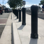 Un trottoir bordé de bornes noires à côté d'une rue ; des gens marchent près d'un front de mer avec une horloge et des arbres en arrière-plan.