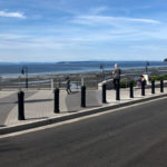 Une promenade en bord de mer avec une tour d'horloge, des gens marchant ou assis sur des bancs, et une vue sur l'océan et la terre lointaine sous un ciel partiellement nuageux.