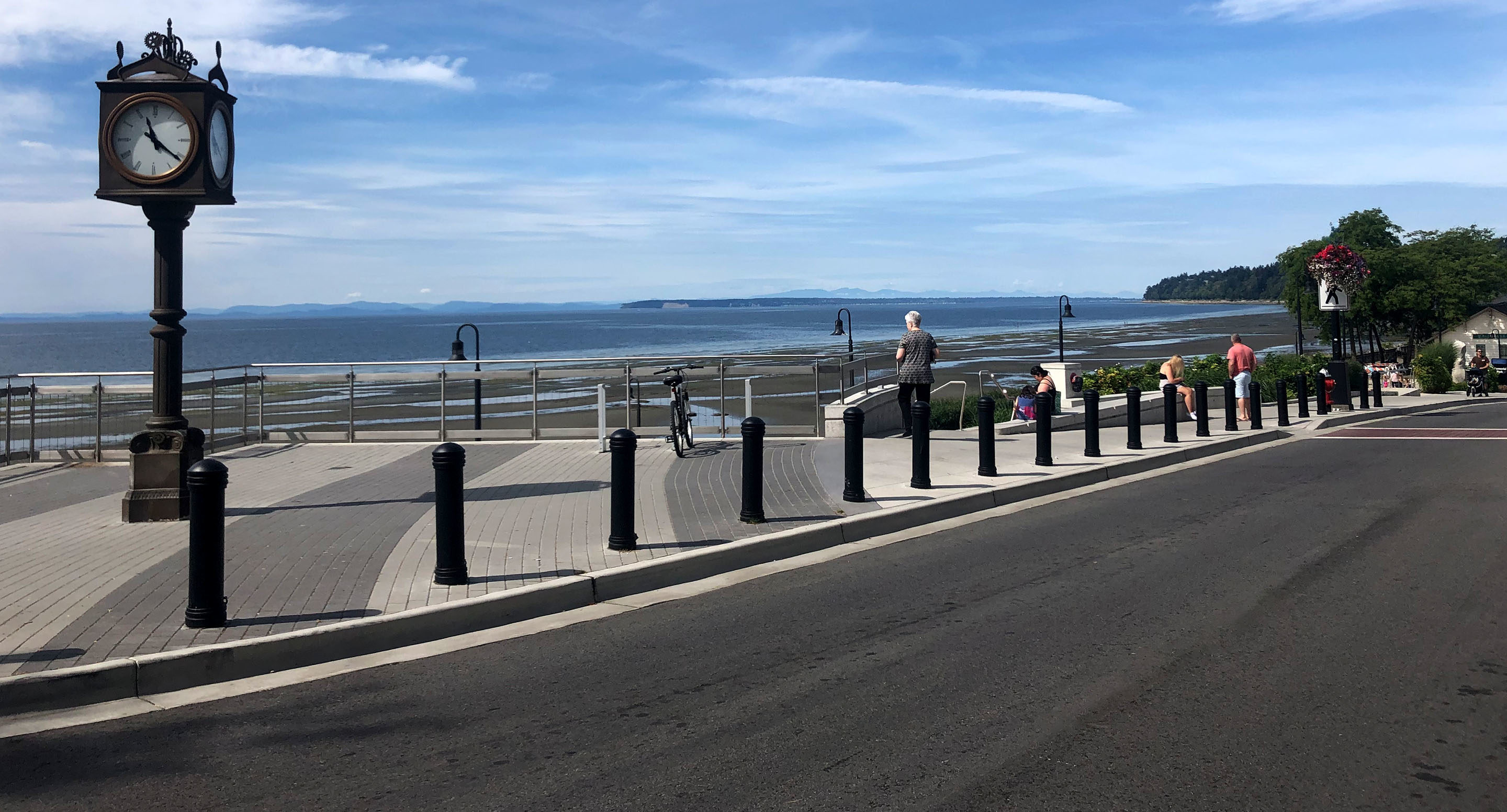 maglin_mbo-0650-bollards-2 Une promenade en bord de mer avec une tour d'horloge, des gens marchant ou assis sur des bancs, et une vue sur l'océan et la terre lointaine sous un ciel partiellement nuageux.