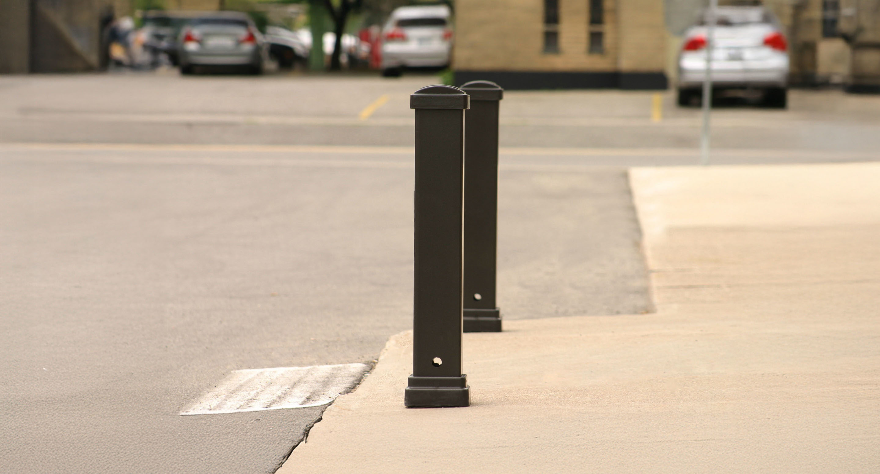 Deux bornes noires se trouvent sur le bord d'un trottoir à côté d'un parking, avec des voitures garées et un bâtiment en arrière-plan.