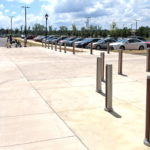 Wide, sunny sidewalk with metal bollards, a parking lot with parked cars, and a few people walking in the distance.