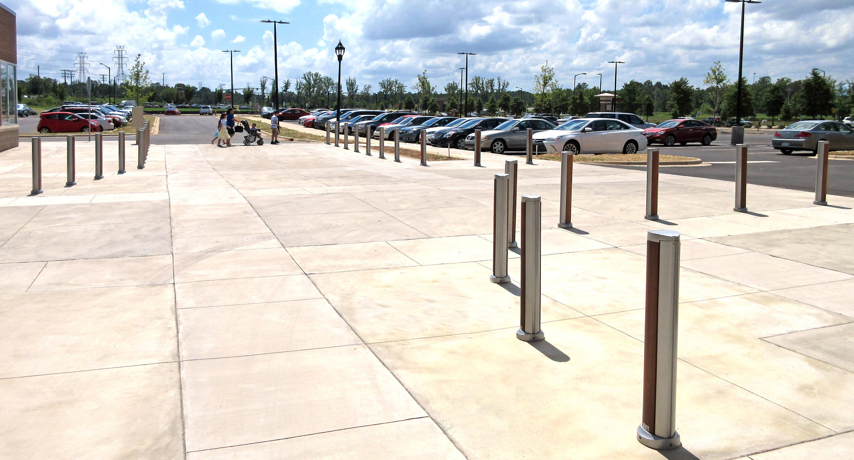 maglin_mbo-1600-bollards-1 Wide, sunny sidewalk with metal bollards, a parking lot with parked cars, and a few people walking in the distance.
