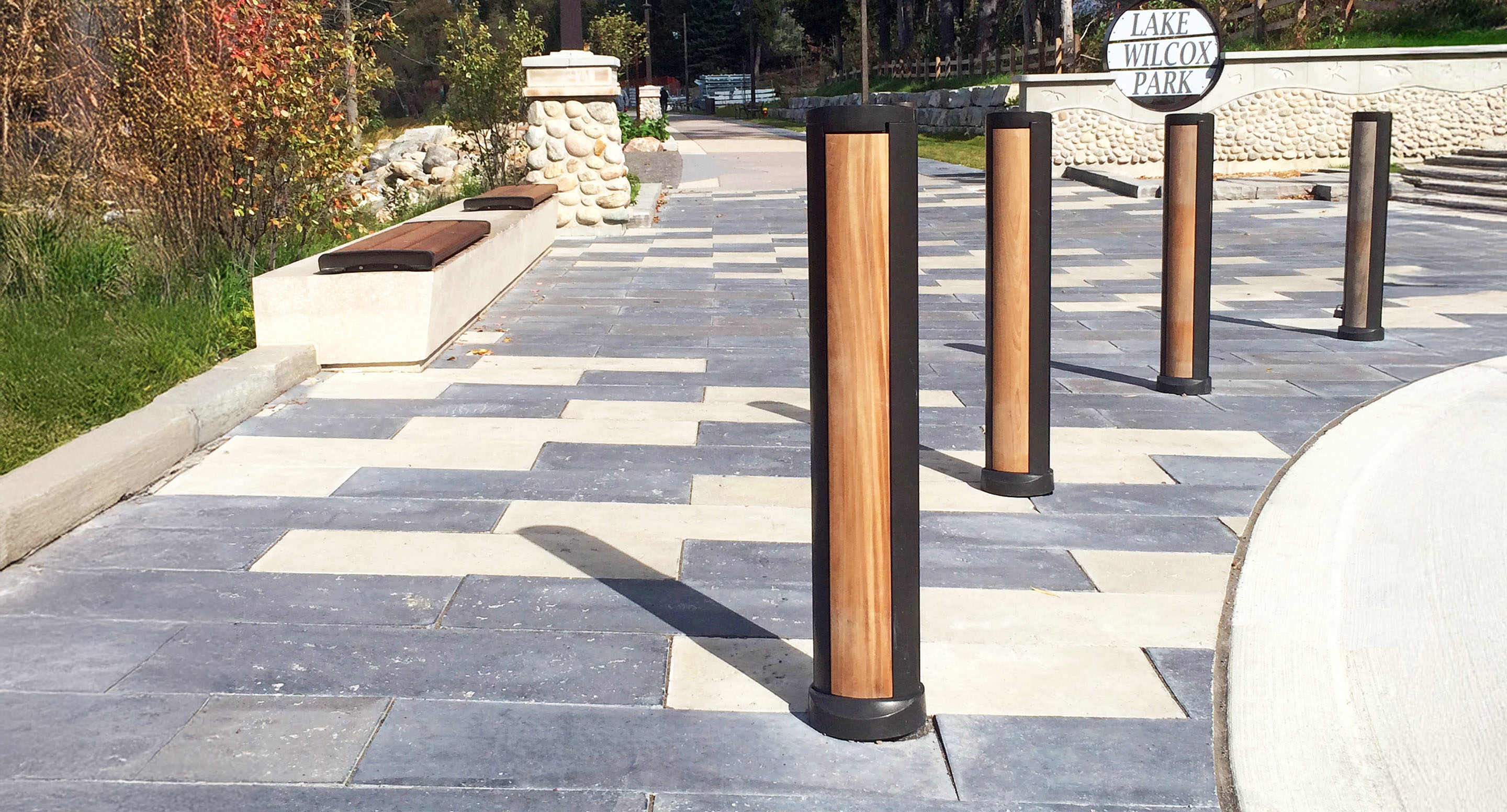 maglin_mbo-1600-bollards-2 Four wooden bollards are installed along a paved pedestrian pathway near a stone bench and the entrance sign for Lake Wilcox Park.