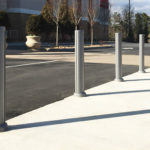 A row of metal bollards lines the edge of a sidewalk next to a parking lot on a clear day.