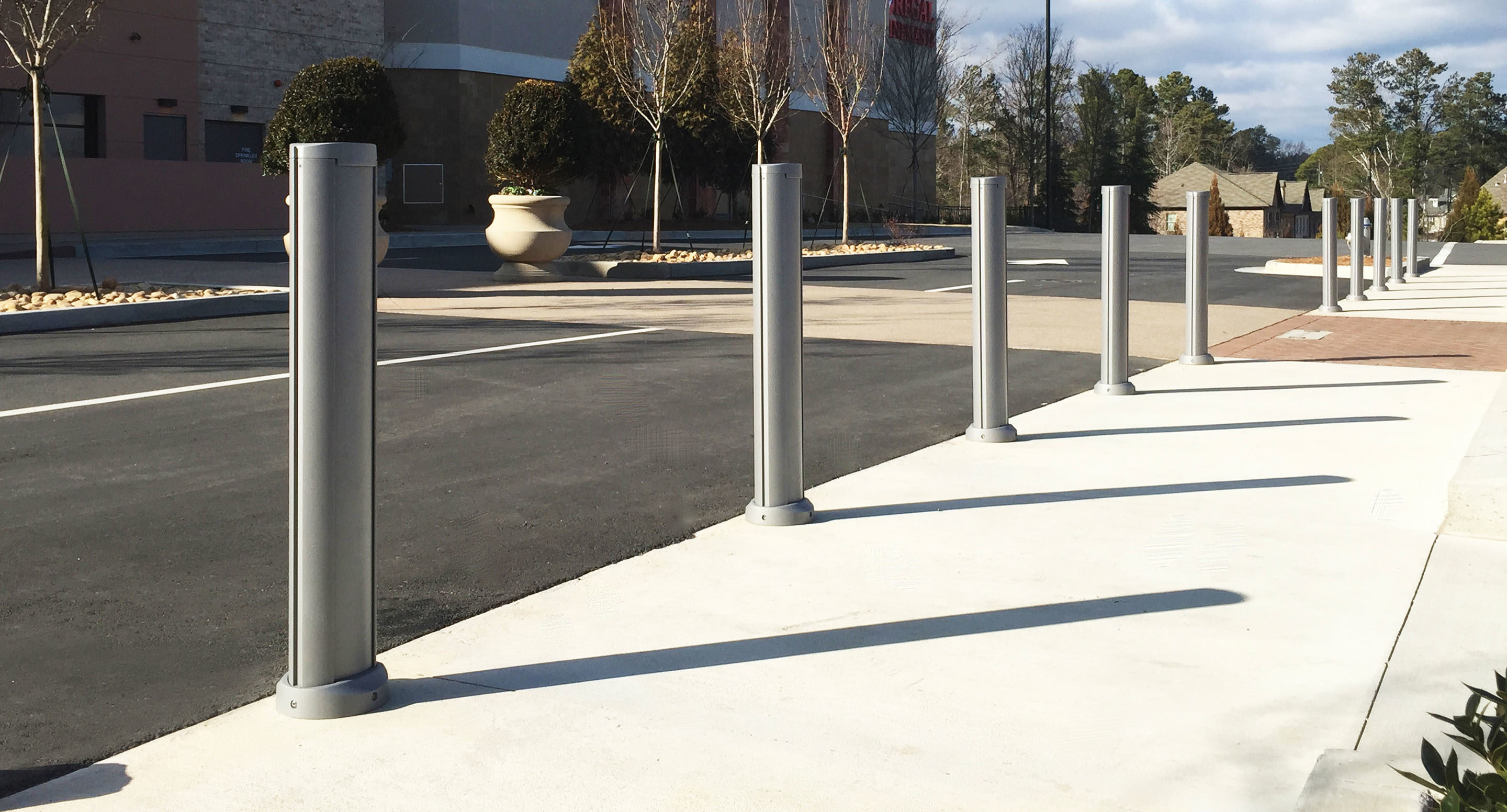 maglin_mbo-1600-bollards-3 A row of metal bollards lines the edge of a sidewalk next to a parking lot on a clear day.