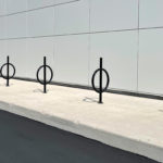 A row of modern black bike racks with circular designs is installed on a concrete sidewalk beside a white tiled building wall.