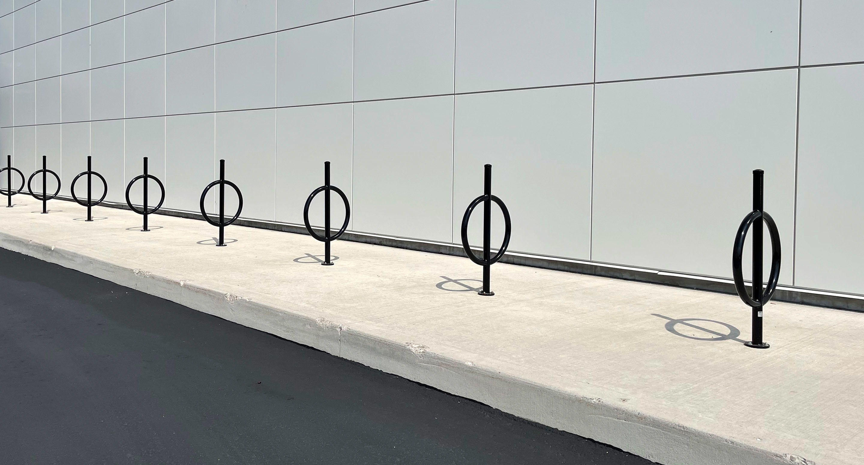 A row of modern black bike racks with circular designs is installed on a concrete sidewalk beside a white tiled building wall.