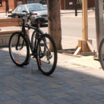 A black bicycle is locked to a metal post on a paved sidewalk near benches and a street with parked cars.