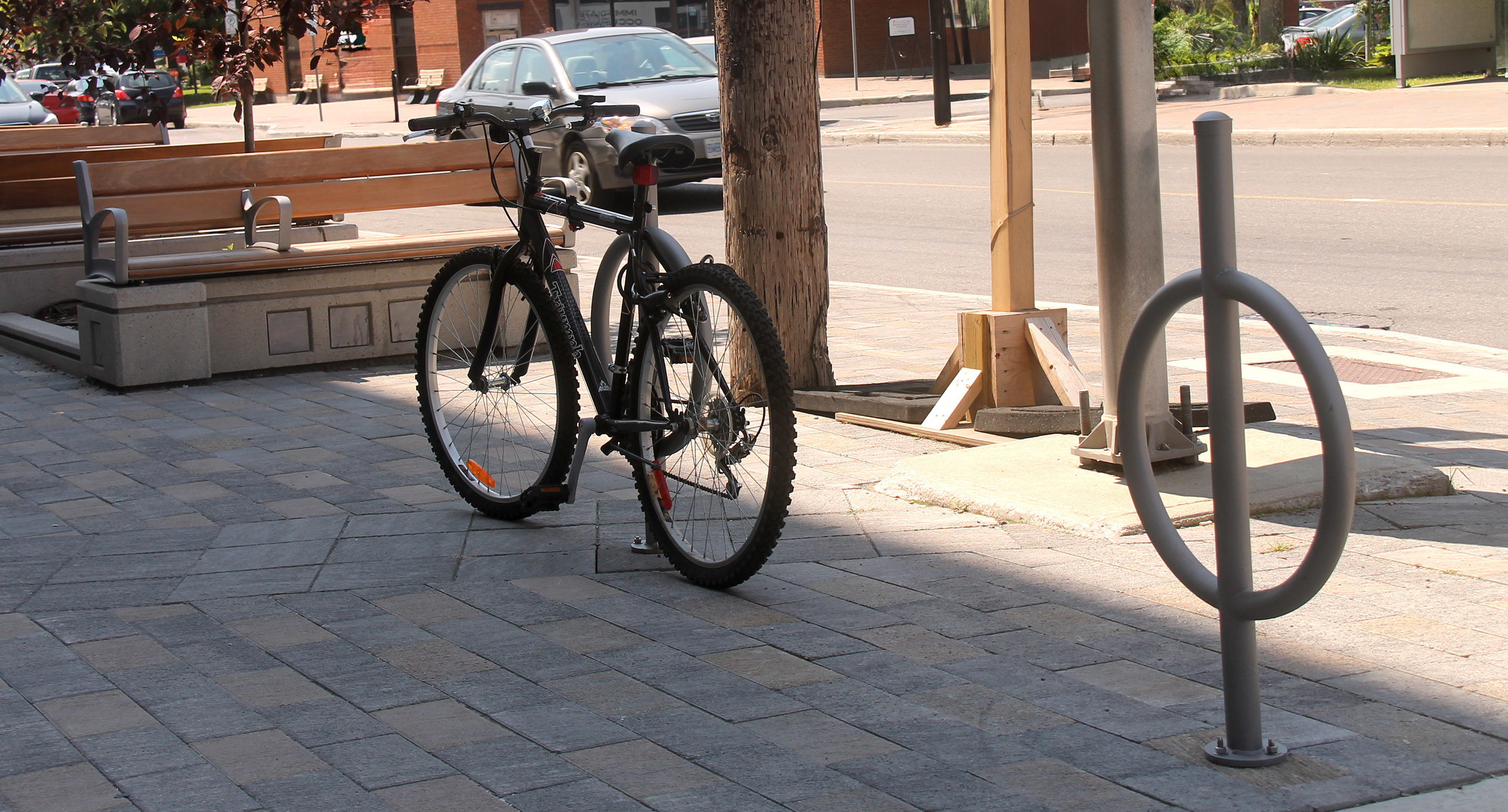 maglin_mbr-0100-bike-racks-2 A black bicycle is locked to a metal post on a paved sidewalk near benches and a street with parked cars.