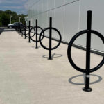 A row of black metal bicycle racks with circular designs is installed on a concrete sidewalk next to a modern building.