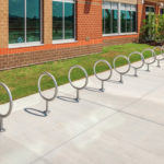 A row of circular metal bike racks installed on a concrete sidewalk outside a brick building with large windows.