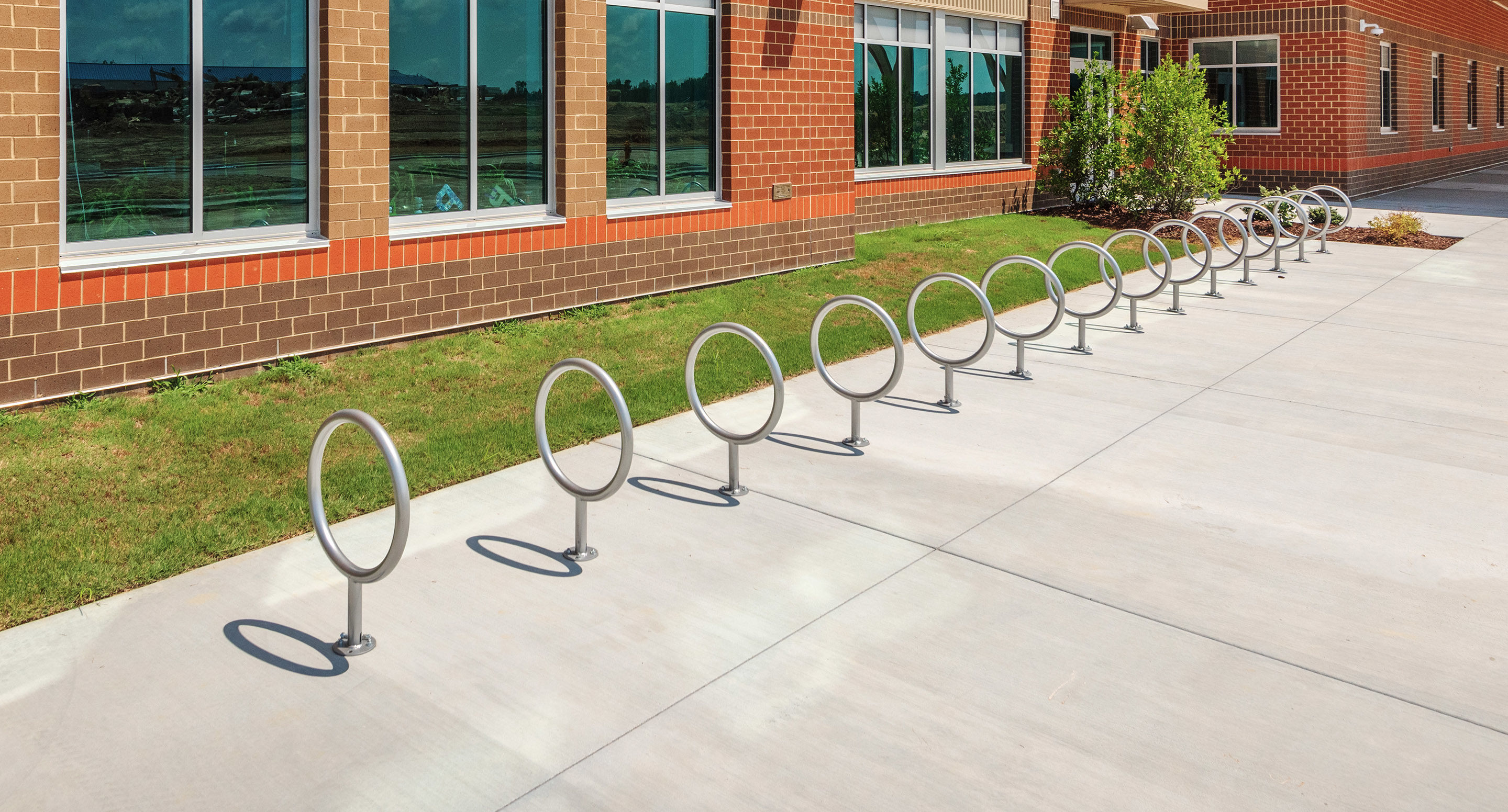 maglin_mbr-0150-bike-racks-1 A row of circular metal bike racks installed on a concrete sidewalk outside a brick building with large windows.