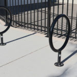 Two black metal bike racks shaped like circles are installed on a light-colored concrete sidewalk near a black metal fence.