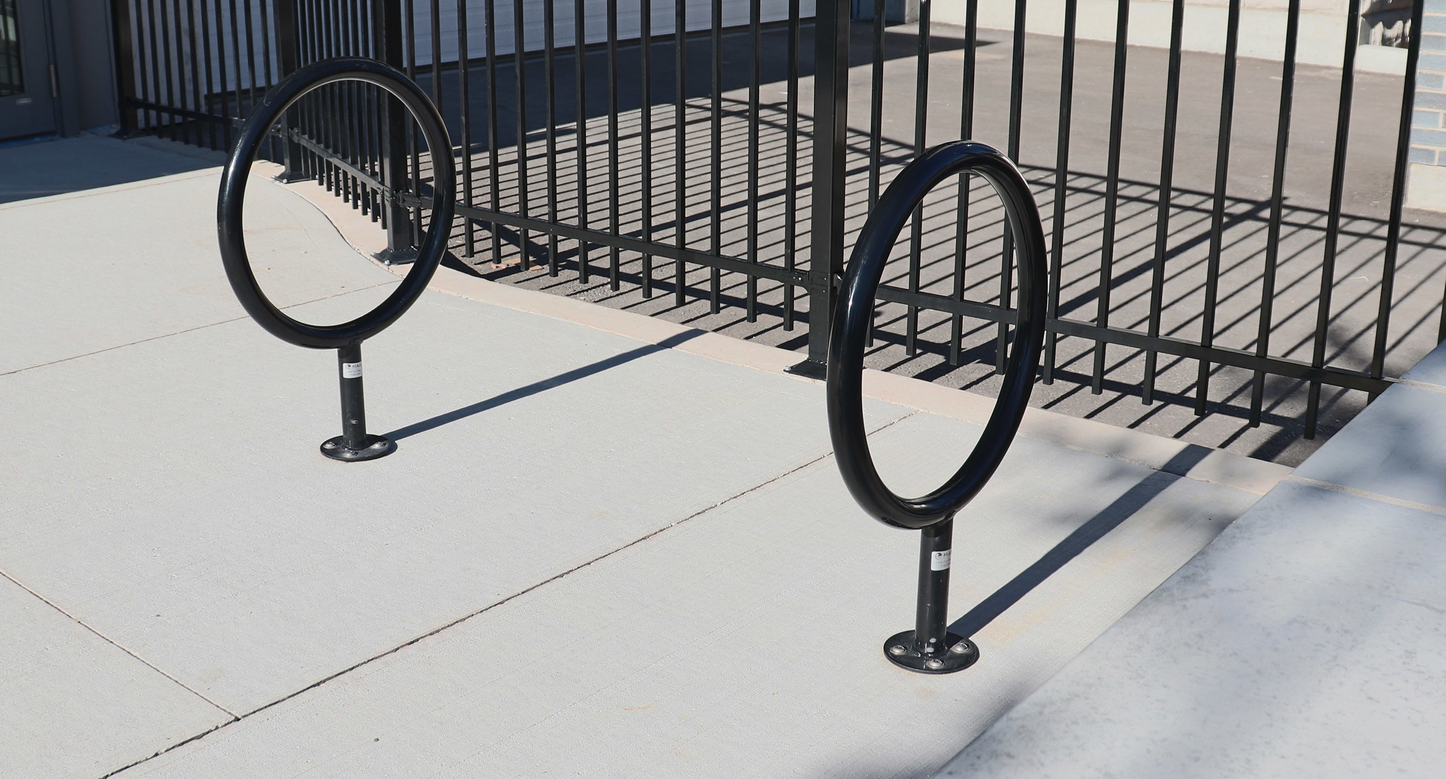 maglin_mbr-0150-bike-racks-2 Two black metal bike racks shaped like circles are installed on a light-colored concrete sidewalk near a black metal fence.