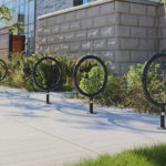 A row of circular metal bike racks is installed on a concrete sidewalk next to a stone building and landscaped greenery.