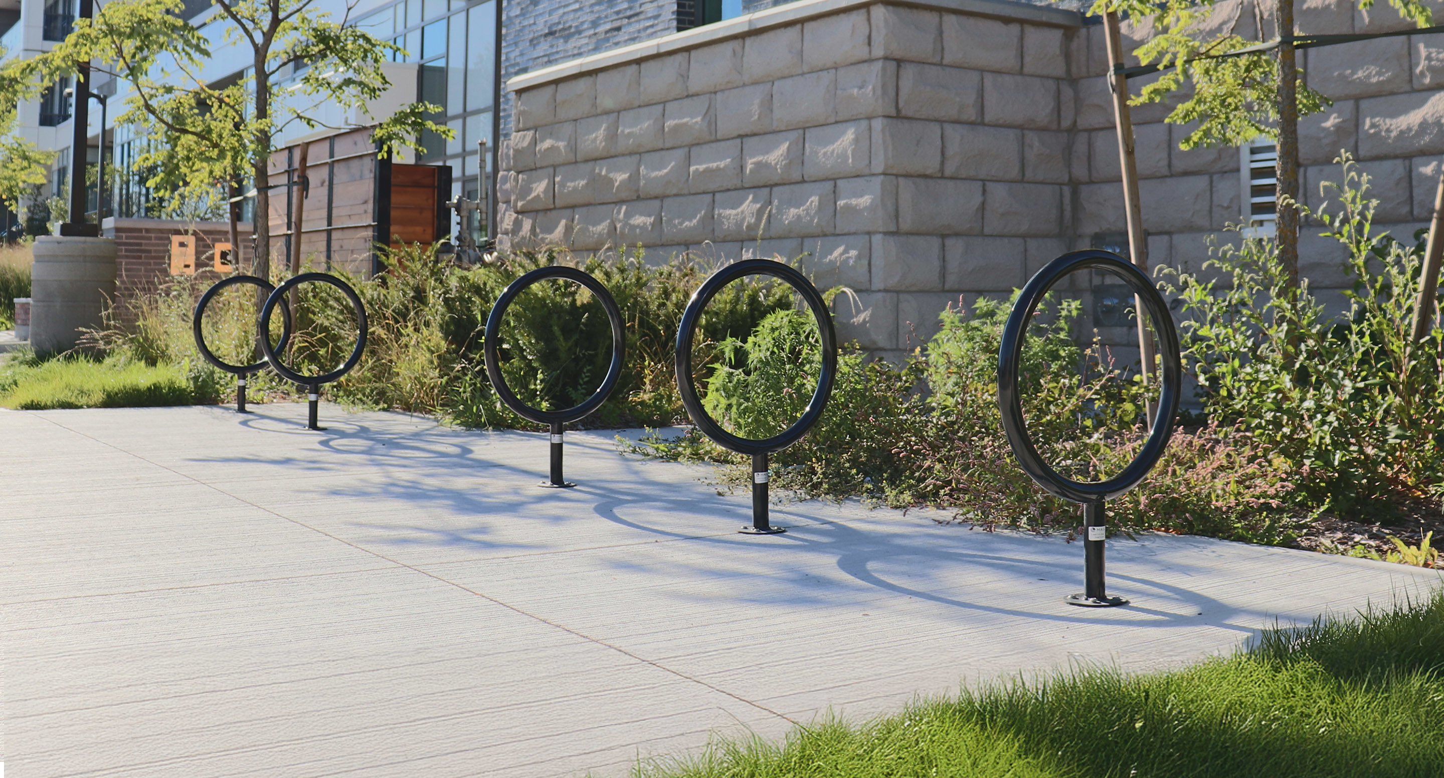 maglin_mbr-0150-bike-racks-3 A row of circular metal bike racks is installed on a concrete sidewalk next to a stone building and landscaped greenery.