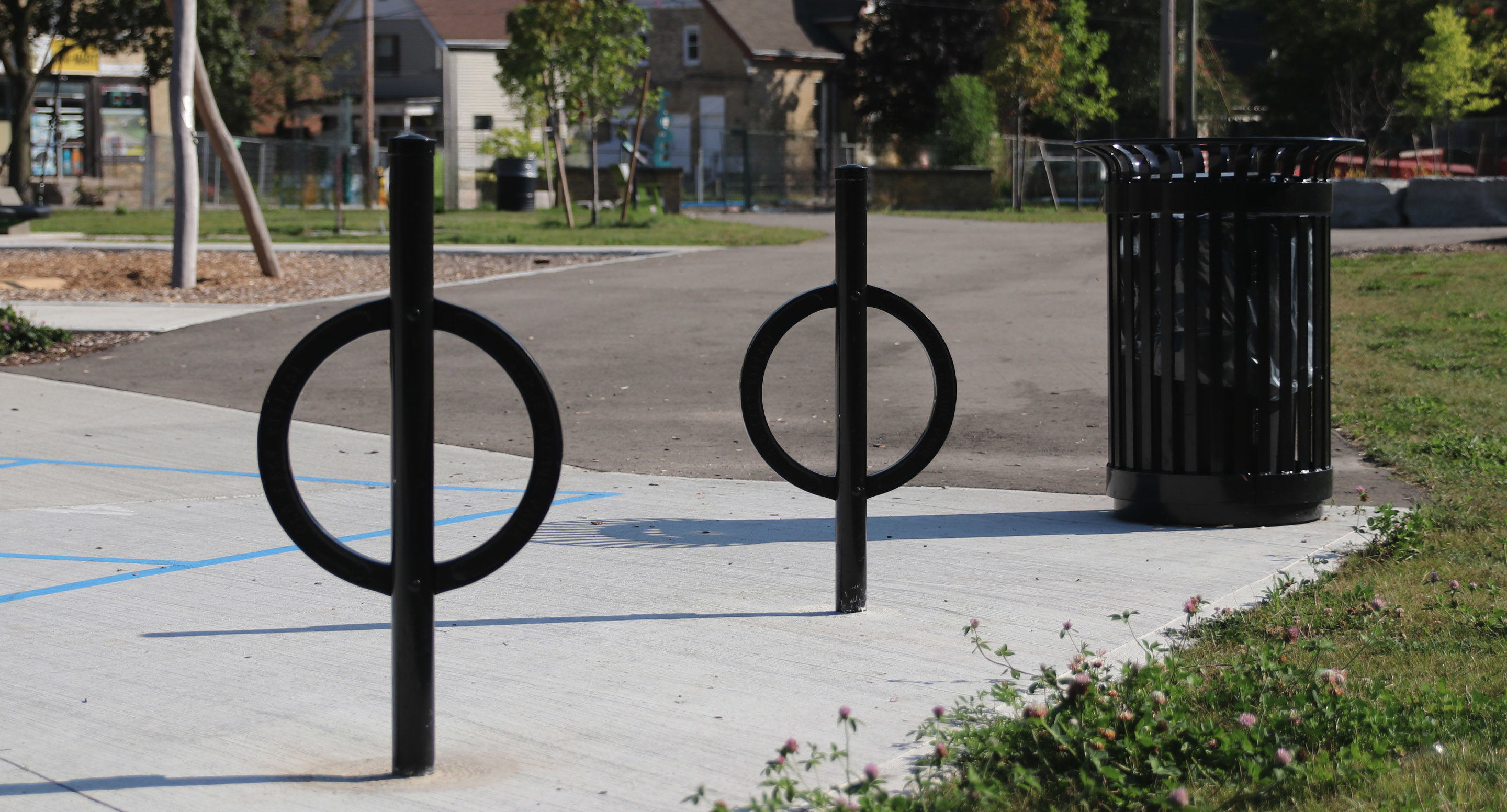 maglin_mbr-0200-bike-racks-1 Deux porte-vélos circulaires noirs et une poubelle noire sont placés sur un trottoir en béton près d'un parc avec de l'herbe, des arbres et un chemin pavé en arrière-plan.