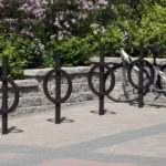 Une bicyclette noire est attachée à un porte-vélos circulaire sur une surface pavée, devant un mur de pierre et des arbustes verts.