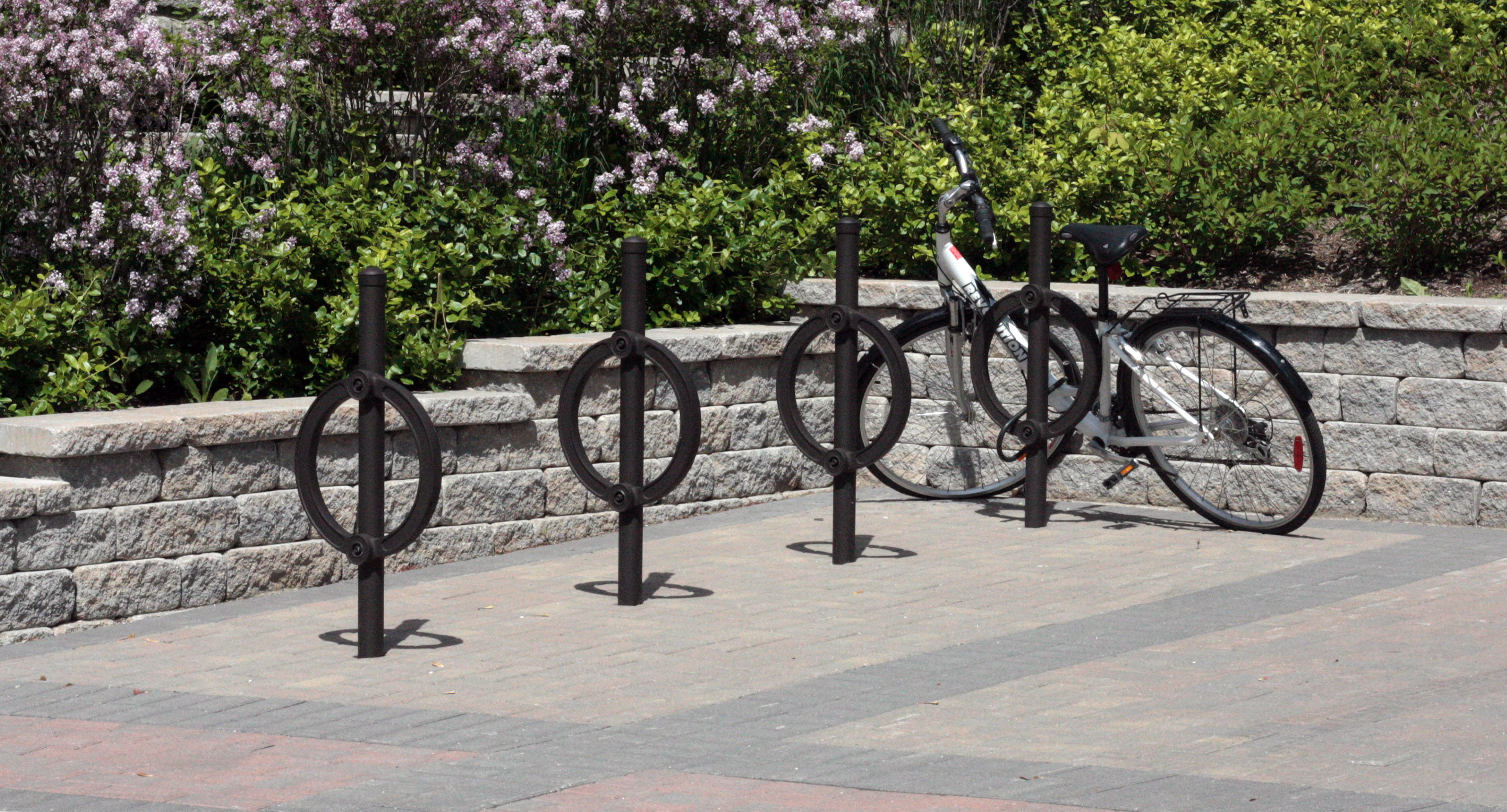 maglin_mbr-0200-bike-racks-2 Une bicyclette noire est attachée à un porte-vélos circulaire sur une surface pavée, devant un mur de pierre et des arbustes verts.