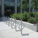 Des supports à vélos en métal sont installés à côté de bancs et d'une jardinière surélevée avec des arbustes et des petits arbres dans un espace urbain extérieur moderne.
