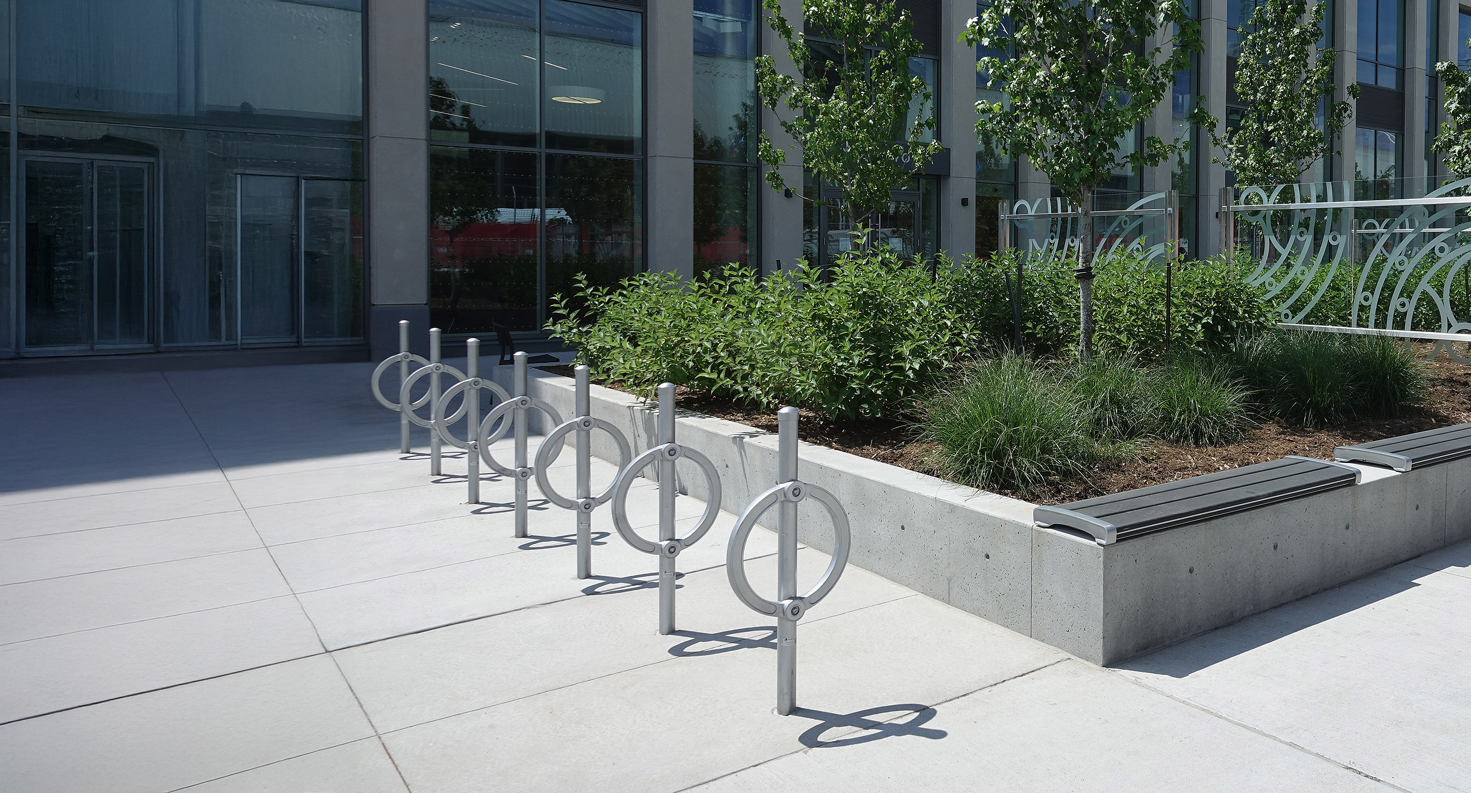 A row of metal bike racks is installed next to a raised planter with green shrubs and benches outside a modern glass building.