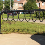 A black metal bike rack with circular slots is installed on a concrete pad in a grassy area near residential houses.