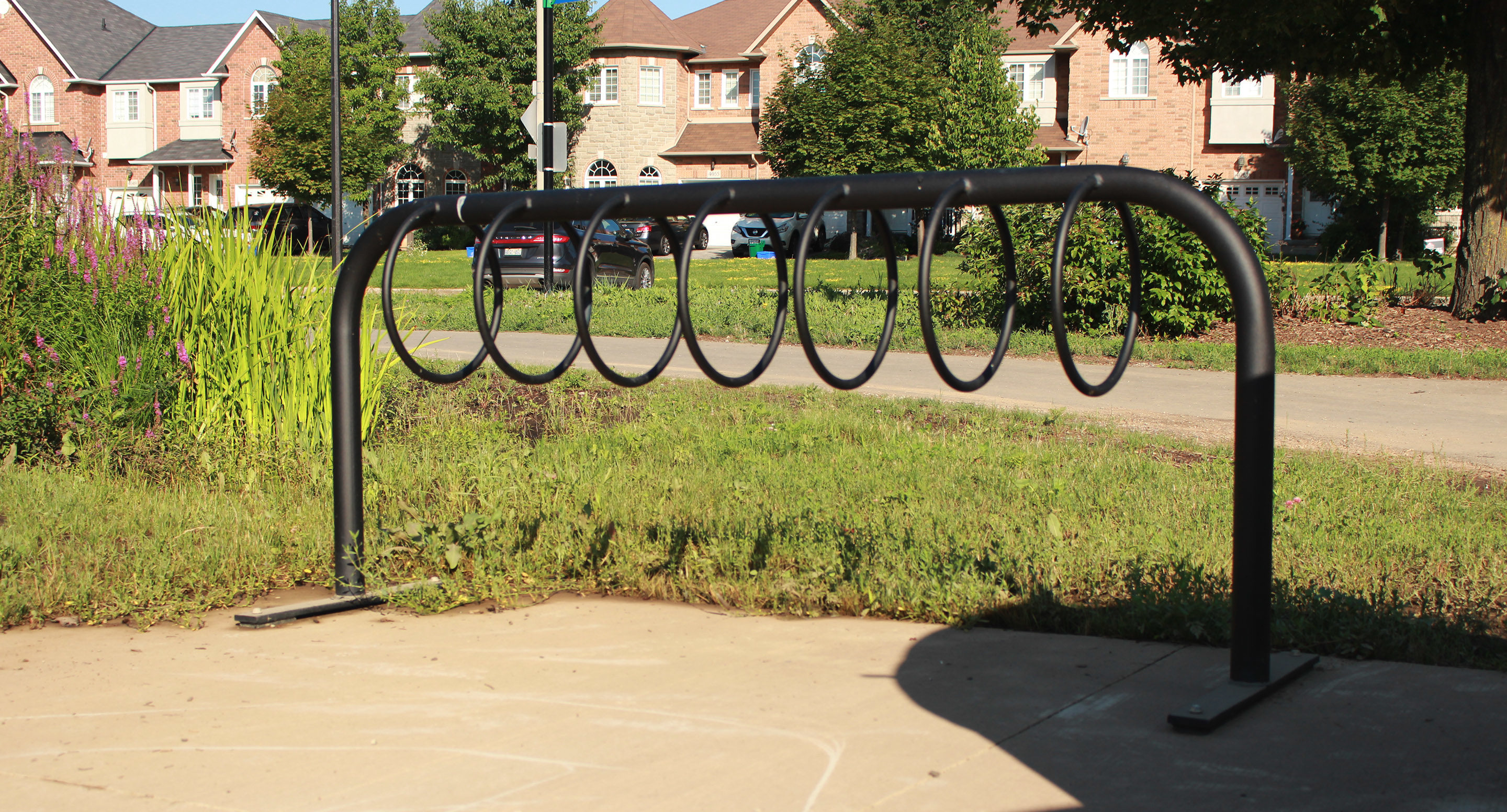 maglin_mbr-0300-bike-racks-1 A black metal bike rack with circular slots is installed on a concrete pad in a grassy area near residential houses.