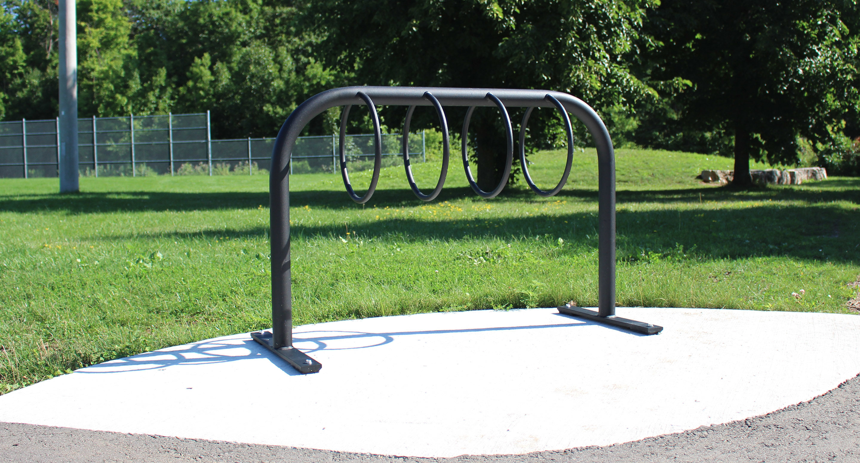 maglin_mbr-0300-bike-racks-2 A black metal bike rack with four loops is installed on a concrete pad in a grassy park area, with trees and a fence in the background.