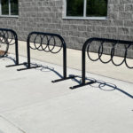 A bike rack with circular slots is installed on a sidewalk near a gray brick building; one bicycle is parked and locked to the rack.